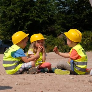 3 Kinder in Warnweste und Bauhelmen sitzen gemütlich auf der Schotterfläche und essen Laugenbrötchen