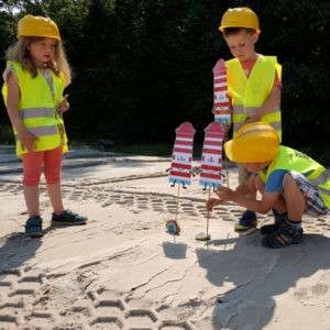 3 Kinder in Warnweste und Bauhelmen stecken gebastelte Leuchttürme in den Sand. 